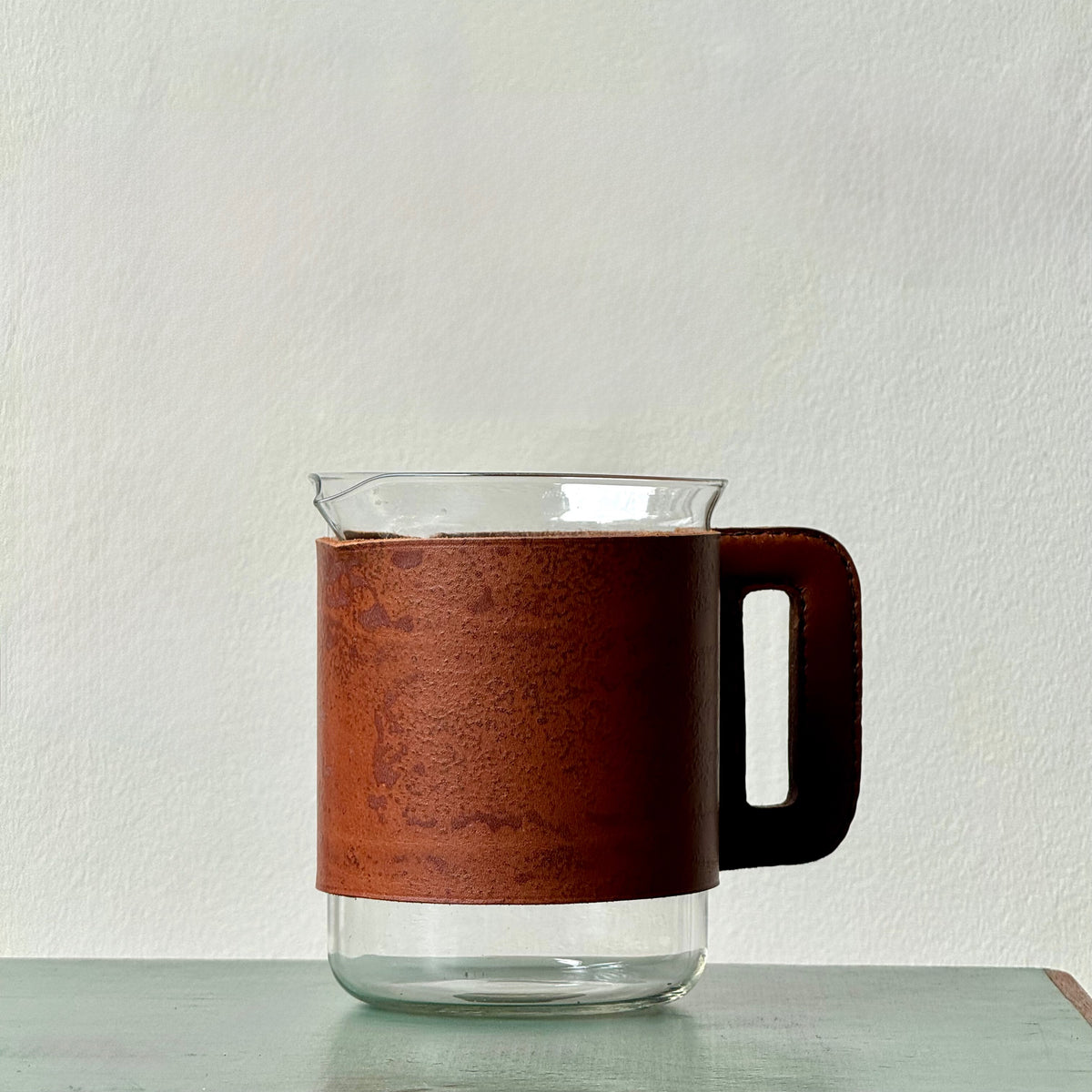 A mouth-blown glass jug with hand stitched leather sleeve handle, made and designed by Austrian designer Carl Auböck II.