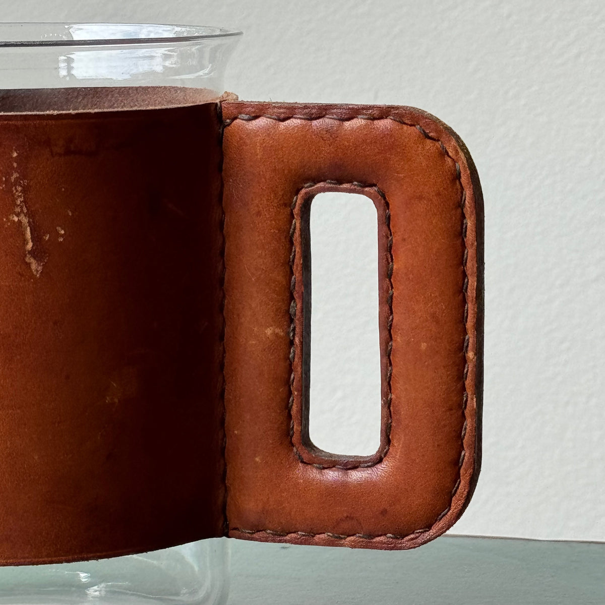 A mouth-blown glass jug with hand stitched leather sleeve handle, made and designed by Austrian designer Carl Auböck II.