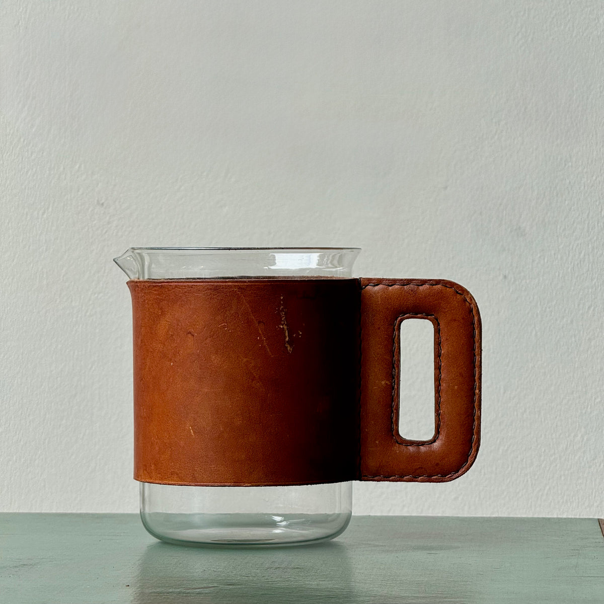 A mouth-blown glass jug with hand stitched leather sleeve handle, made and designed by Austrian designer Carl Auböck II.