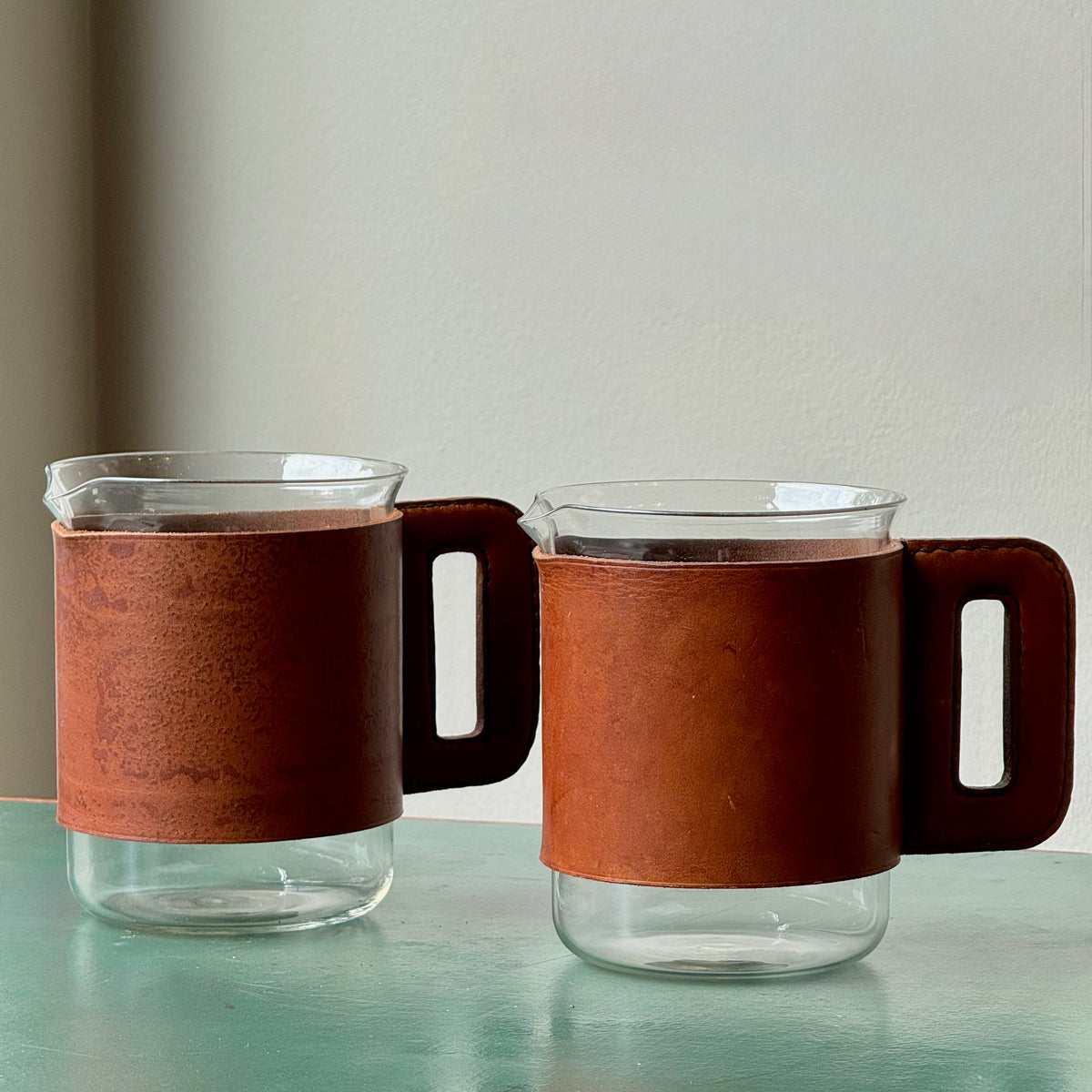 A mouth-blown glass jug with hand stitched leather sleeve handle, made and designed by Austrian designer Carl Auböck II.