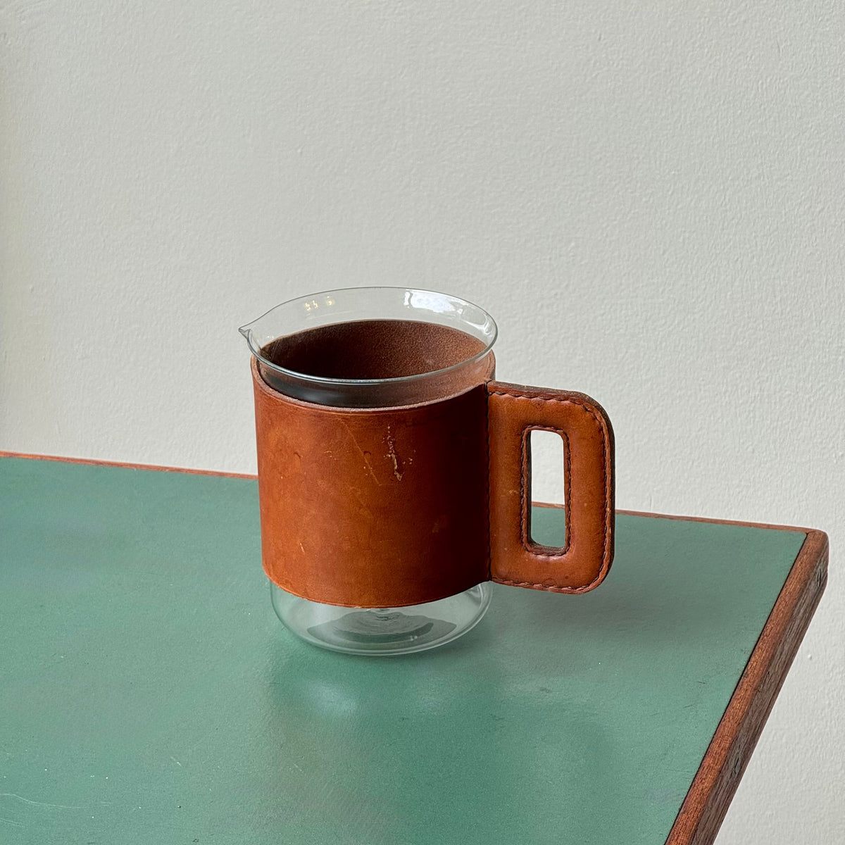 A mouth-blown glass jug with hand stitched leather sleeve handle, made and designed by Austrian designer Carl Auböck II.