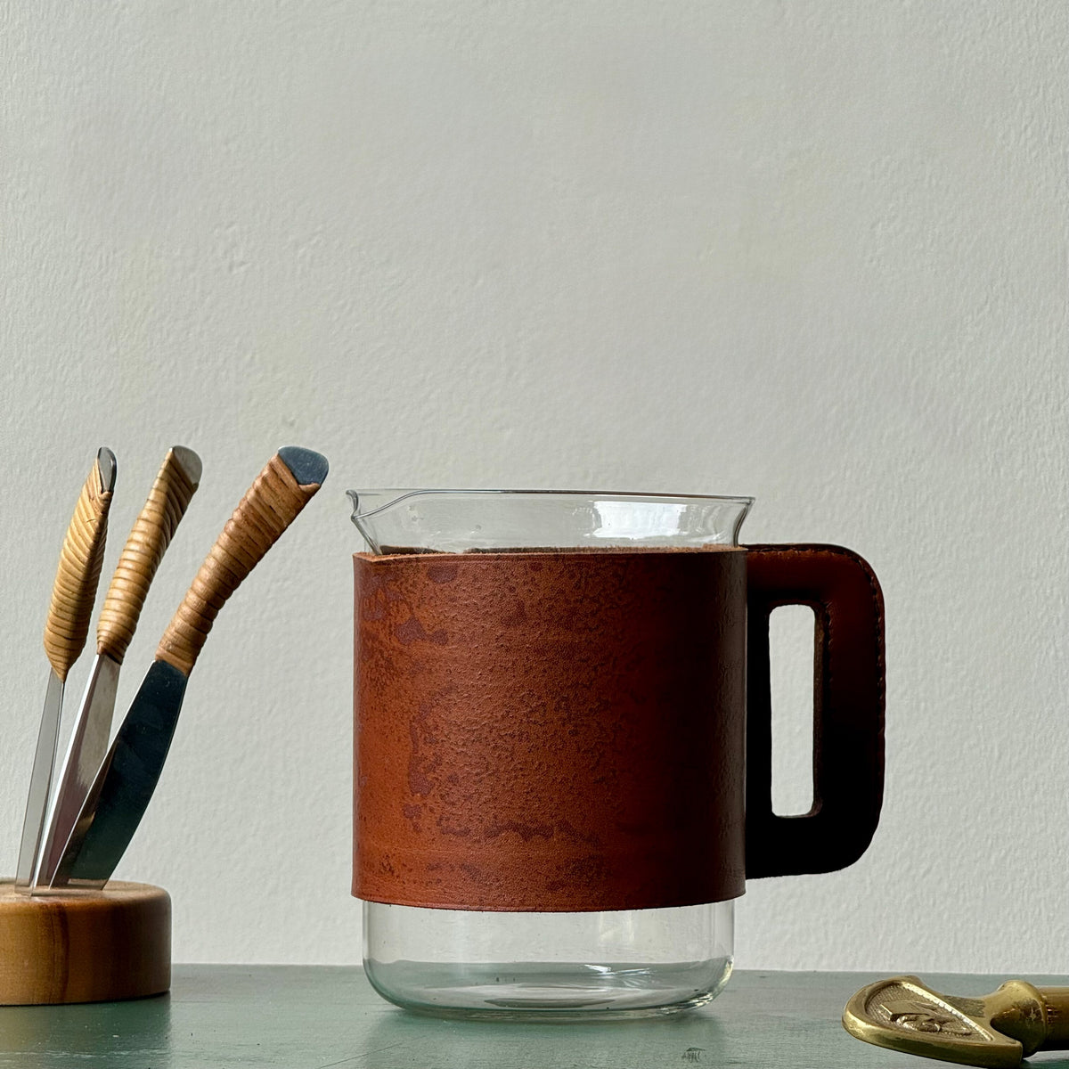 A mouth-blown glass jug with hand stitched leather sleeve handle, made and designed by Austrian designer Carl Auböck II.